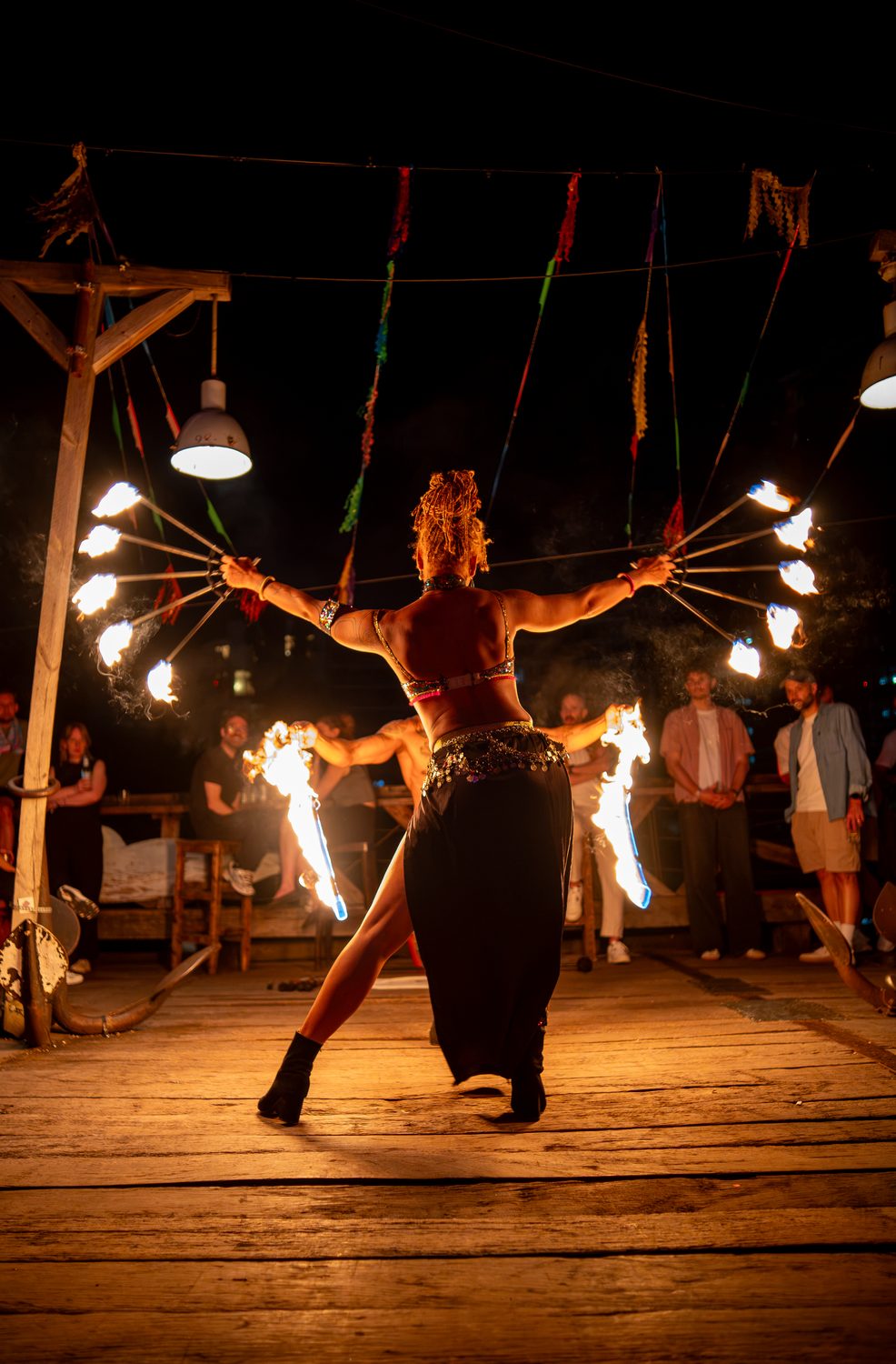 Fire-fan performance seen from behind, flames spread wide before a live audience